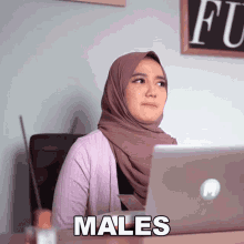 a woman wearing a hijab sits at a desk with a laptop and the word males written on the screen