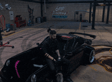 a man sits on the hood of a car in a garage with graffiti on the wall that says 666