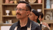 a man with glasses and a mustache stands in front of a shelf that says the great canadian baking show