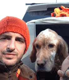 a man wearing an orange beanie stands next to a dog