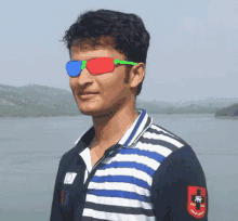 a man wearing a striped shirt and 3d glasses stands in front of a body of water