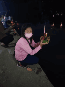 a girl wearing a mask is kneeling down holding a bowl of food in front of a body of water