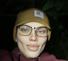 a woman wearing glasses and a carhartt hat makes a funny face