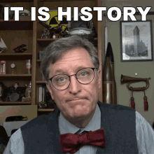 a man wearing glasses and a bow tie is standing in front of a sign that says it is history