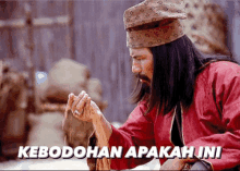 a man with long hair and a beard is holding a piece of wood with the words kebodohan apakah ini written below him