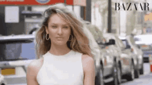 a woman wearing a white dress and hoop earrings is standing in front of a row of cars in a city street .