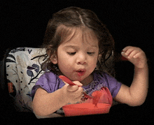 a little girl in a purple shirt is eating from a red bowl with a spoon