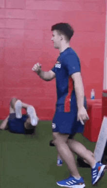 a man in a blue shirt and blue shorts is running in a gym while another man is laying on the floor .