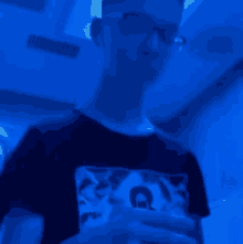 a young man wearing glasses and a black t-shirt is standing in a dark room with a blue light behind him .