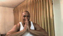 a man in a white tank top is smiling with his hands folded in front of a curtain