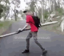 a man in a red shirt is walking down a road with a backpack and holding a large stick .