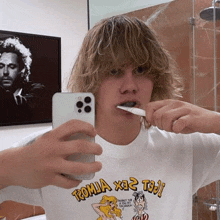 a young man is brushing his teeth in a bathroom while taking a selfie with his cell phone .