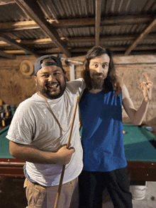 two men are posing for a picture in front of a pool table with one wearing a blue tank top