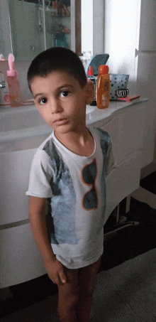 a young boy with sunglasses on his shirt stands in front of a bathroom counter