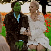 two women in costumes are sitting in a field of flowers . one of the women is green and the other is blonde .