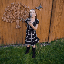 a little girl in a black and white dress stands in front of a wooden fence with a tree on it