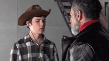 a man with a beard wearing a cowboy hat talks to another man in a plaid shirt