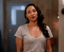 a woman in a grey t-shirt and hoop earrings is standing in a hallway .