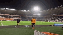 a man in an orange shirt stands on a soccer field with a sign that says train like a pro