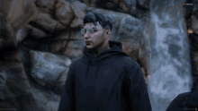 a man wearing sunglasses and a black hoodie stands in front of a waterfall in a video game