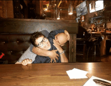 a man is hugging another man while sitting at a table in front of a bar