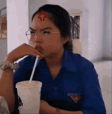 a woman wearing glasses and a blue shirt has a red star on her forehead while drinking from a cup