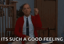 a man in a red sweater and tie is sitting in a chair giving a thumbs up .