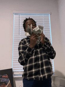 a man in a plaid shirt holds a stack of money in front of a window