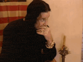 a man with long black hair is sitting at a table with his hand to his mouth .