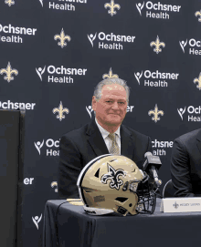 a man sitting at a table with a football helmet in front of a ochsner health backdrop