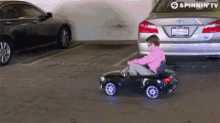 a child is driving a toy car in a parking lot with a spinnin ' tv logo in the background