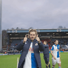 a man giving a thumbs up on a soccer field wearing a jacket that says ' x1 ' on it