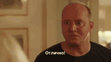 a bald man in a black shirt is making a funny face and saying " отлично " in a foreign language .