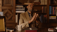 a man sits at a desk in front of a bookshelf with a mug that says i 'm a scientist