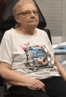 an elderly woman is sitting in a chair wearing a white shirt with a picture of butterflies on it .