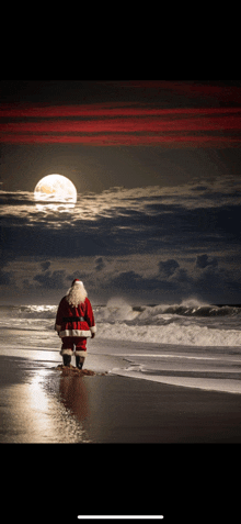 a man in a santa suit stands on a beach looking at the full moon