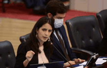 a woman speaking into a microphone while a man wearing a mask looks on