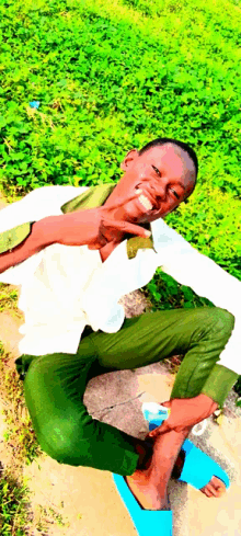 a young man in a white shirt and green pants is sitting on the ground with his legs crossed and smiling