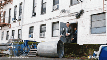 a man in a suit and tie is standing in front of a building with a trash can in front of it and a nbc logo