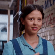 a woman in a blue shirt and blue overalls is standing in front of a brick wall with the word milk on it .