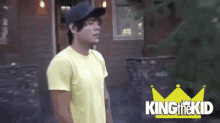 a man wearing a yellow shirt and a blue hat is standing in front of a house with the words king the kid on the bottom