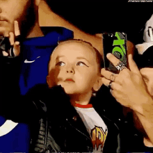 a little girl in a leather jacket is looking up at a man holding a cell phone in a crowd .