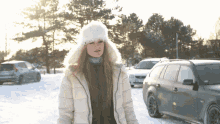 a woman wearing a white hat and a white jacket stands in the snow in front of a row of cars