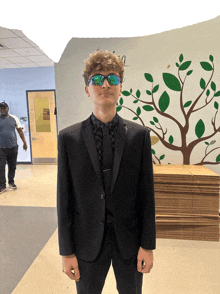 a man in a tuxedo and sunglasses stands in front of a wall with a tree painted on it