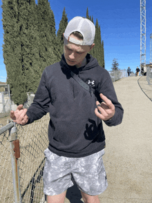 a man wearing a black under armour hoodie is standing next to a chain link fence