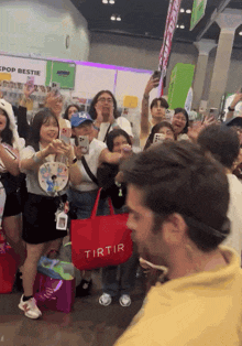 a man in a yellow shirt stands in front of a crowd of people with a red tote bag that says tirtir