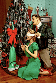 a man blindfolds a woman in front of a christmas tree with a green vacuum cleaner