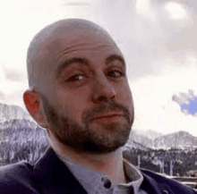 a bald man with a beard is wearing a suit and looking at the camera with mountains in the background .