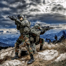 two soldiers are standing on top of a rocky hill aiming their guns at something