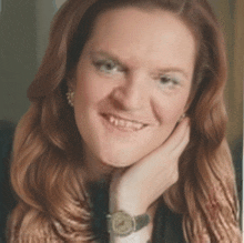 a woman wearing a watch and earrings smiles with her hand on her chin